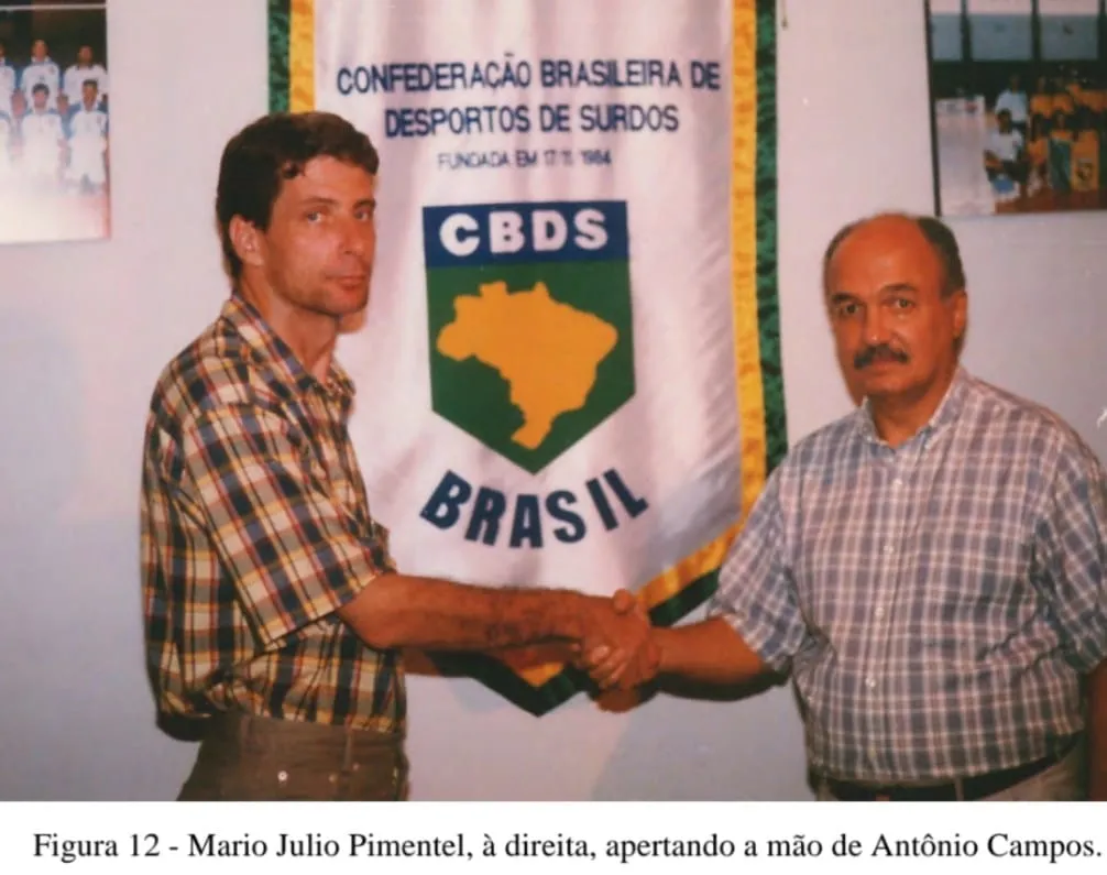 Antônio Campos de Abreu cumprimenta Mario Julio Pimentel diante do estandarte da Confederação Brasileira de Desportos de Surdos.