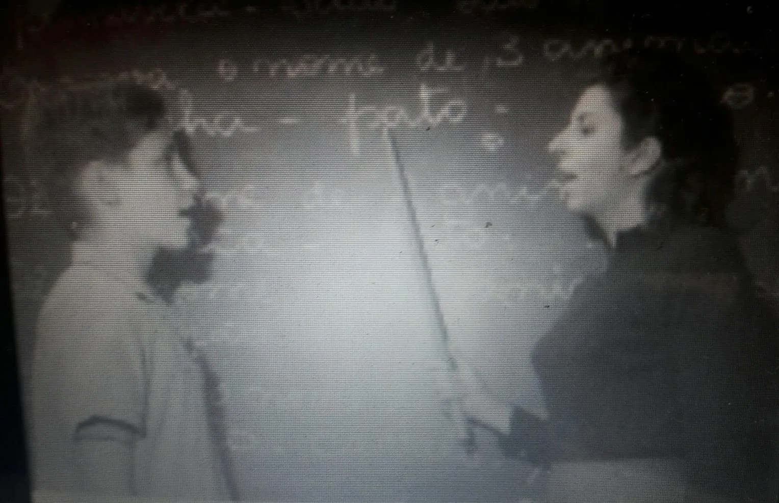 Professora com ponteiro diante do quadro-negro conduzindo exercício oral com um menino em sala de aula.