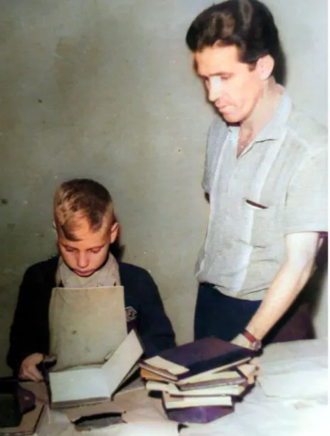 Menino de uniforme escolar folheia um livro ao lado de um adulto, junto a pilhas de livros sobre a mesa.