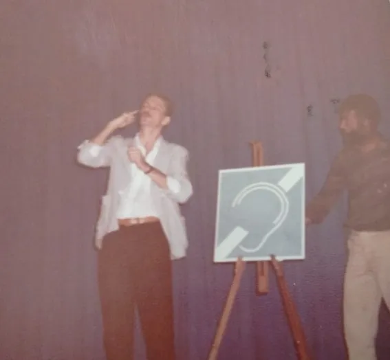 Homem sinalizando em um palco ao lado de painel com o símbolo internacional da surdez.