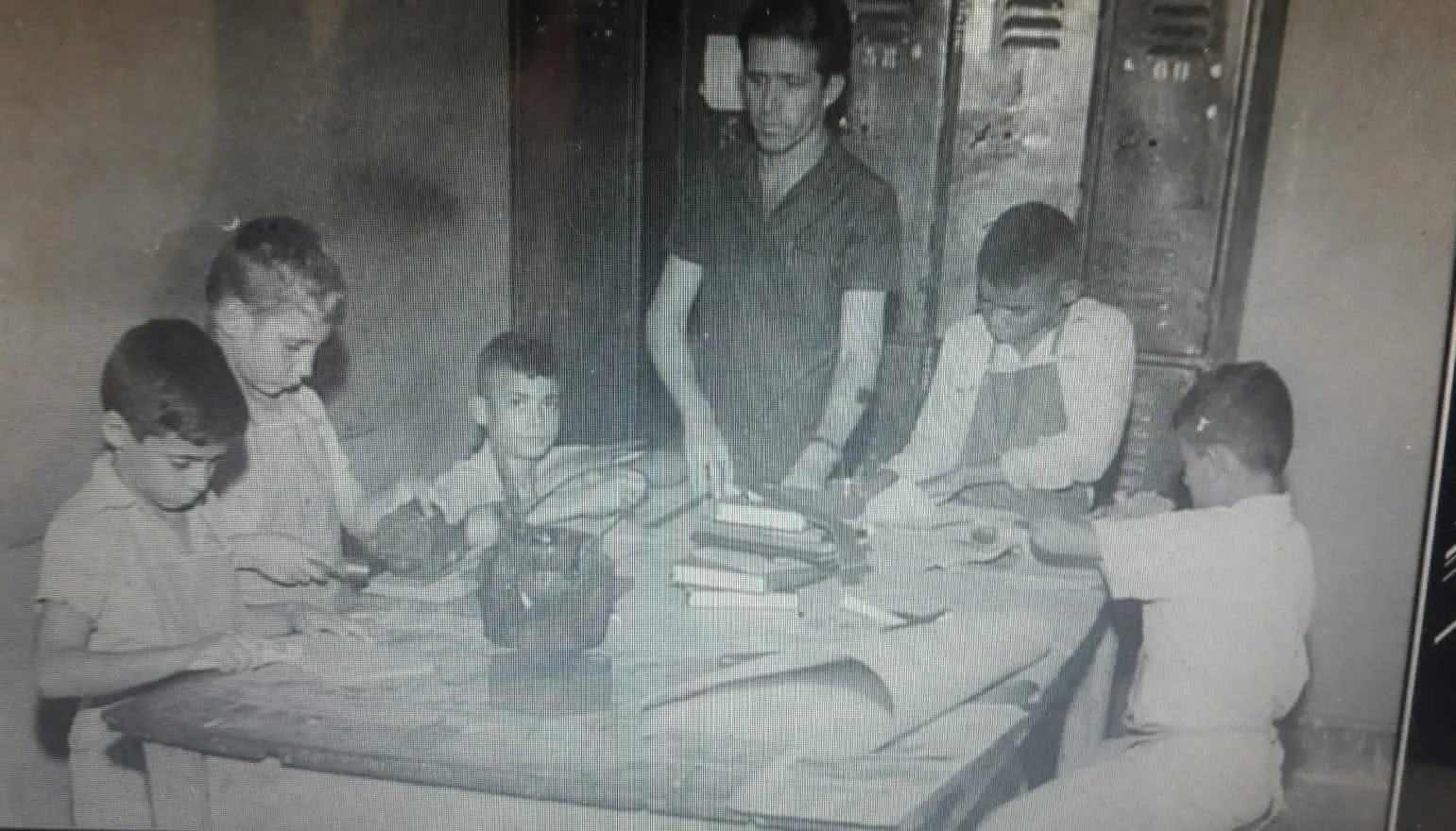 Meninos trabalhando com livros e ferramentas em uma oficina escolar, acompanhados por um instrutor.